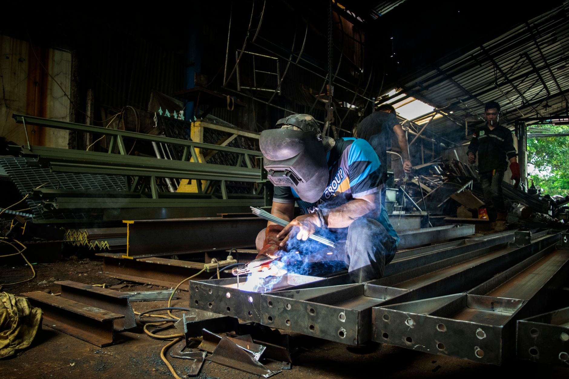 Welder working in a fabrication shop with steel assemblies and bright arc light in the background.