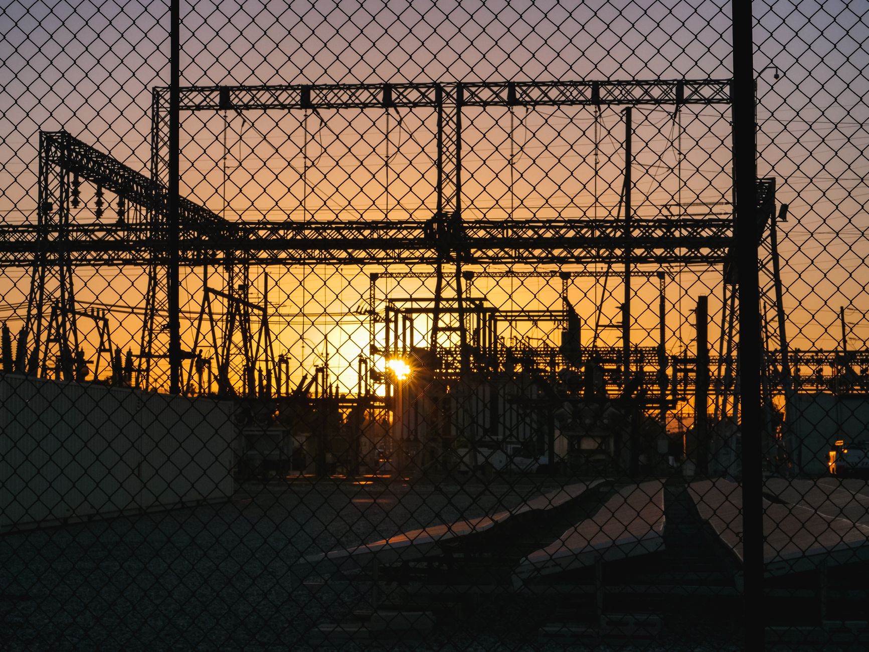 A wide shot of an electrical transmission substation at dusk, its complex grid of wires and transformers silhouetted against the sky, representing a critical infrastructure site.
