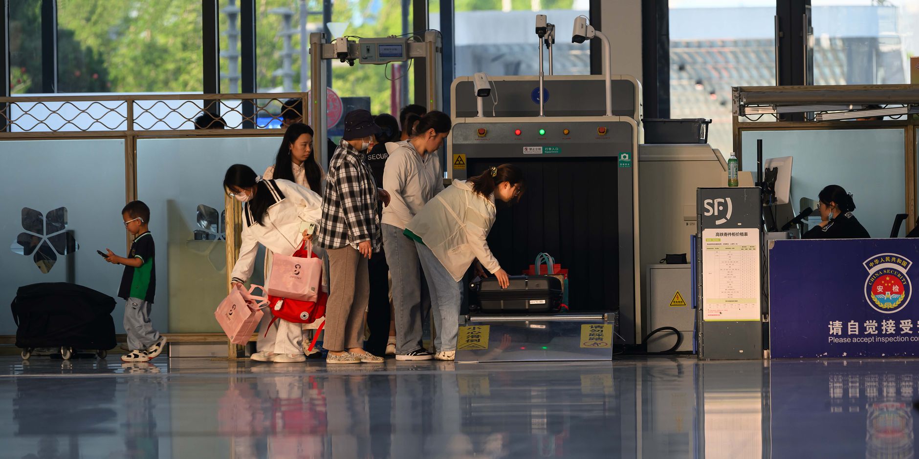 Travelers move through a busy airport security checkpoint with queue lanes and screening stations visible.