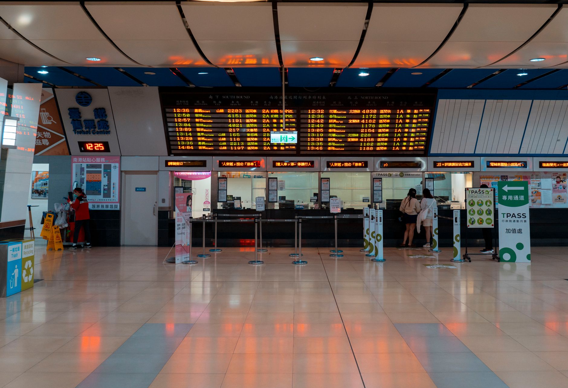 Visitors wait in a busy public-service lobby with queue lanes, fixed seating, and service counters.