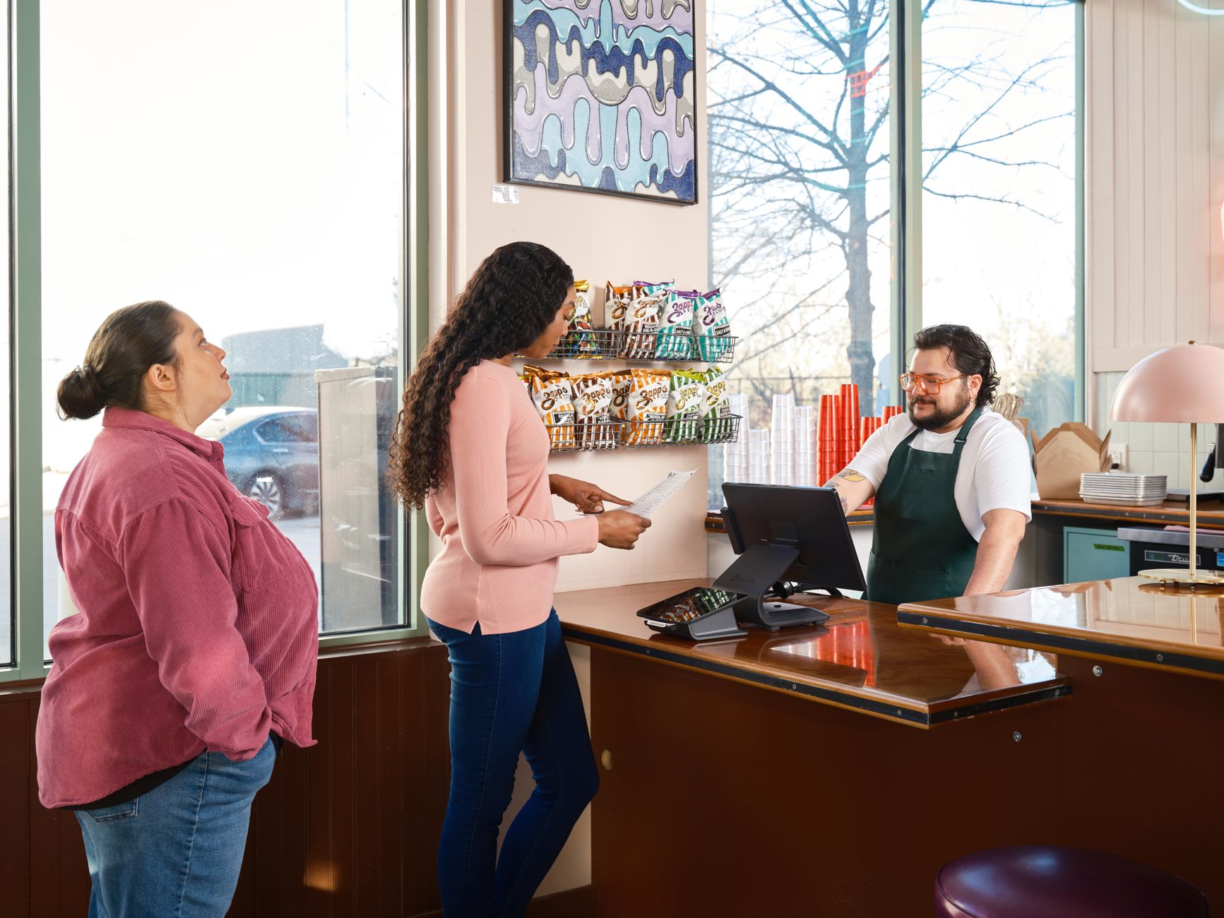 A busy trade counter with staff helping a customer while back-room fulfillment pressure builds during the morning rush.