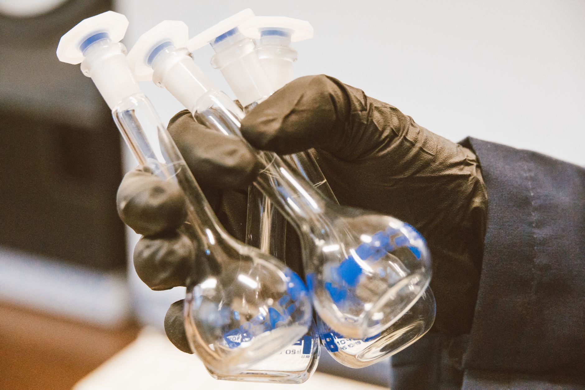 Gloved hands carefully hold a lens by its edge to show the kind of delicate handling needed before polishing and inspection.
