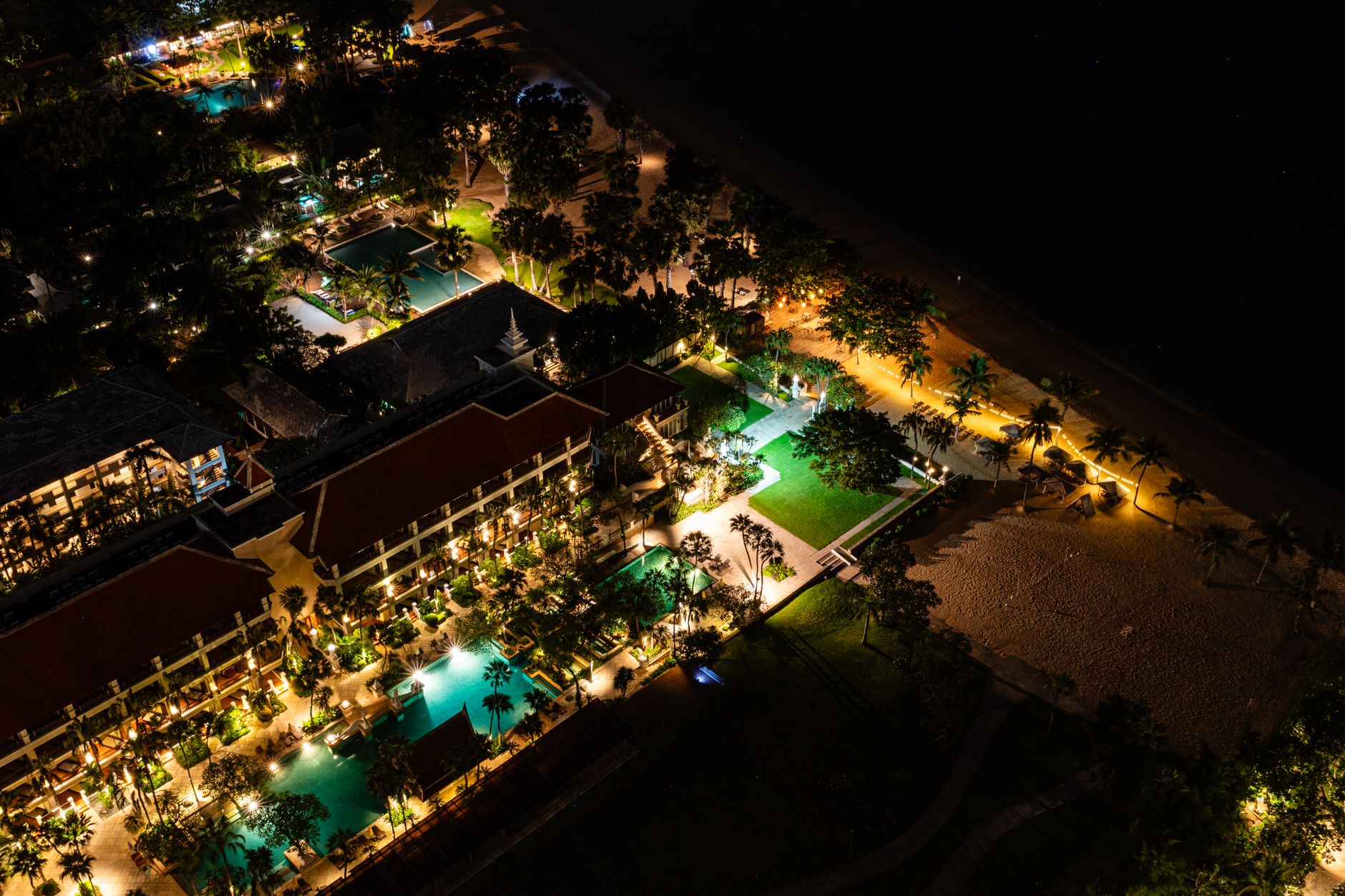 A beachfront resort glows after dark beside a quiet shoreline and access paths.