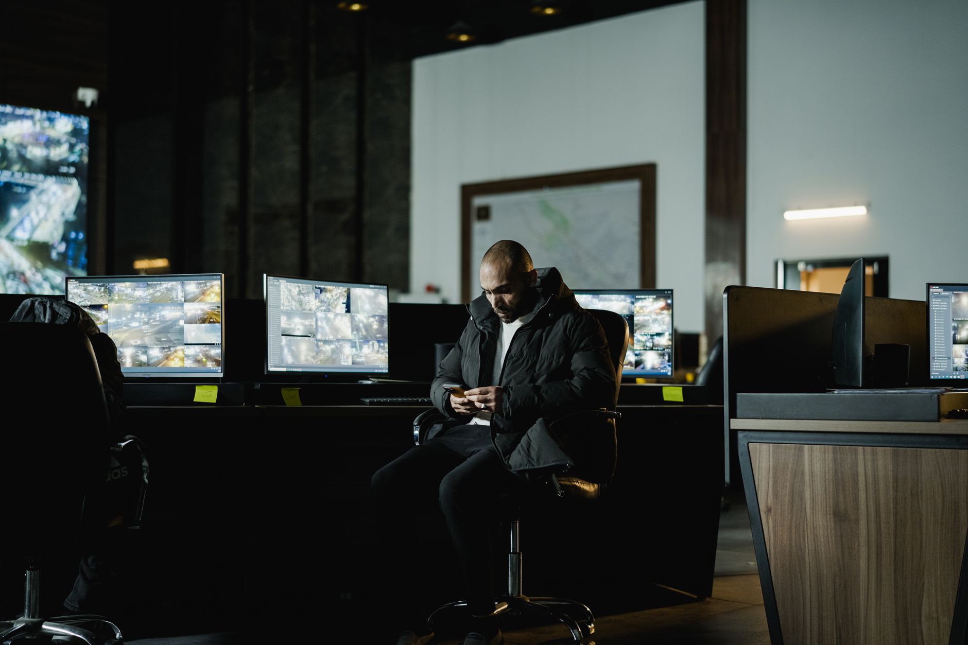 A security guard sits in a dark control room, observing multiple computer monitors displaying surveillance feeds.