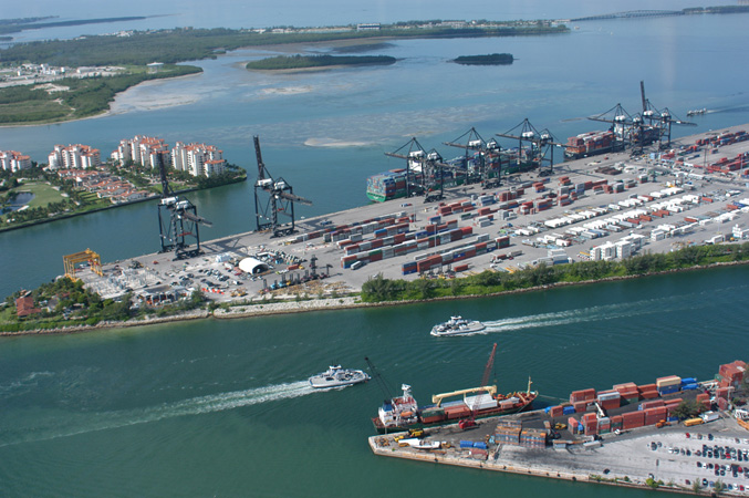 An aerial view of PortMiami showing cruise ships, terminals, and the long passenger concourses of the busy seaport.