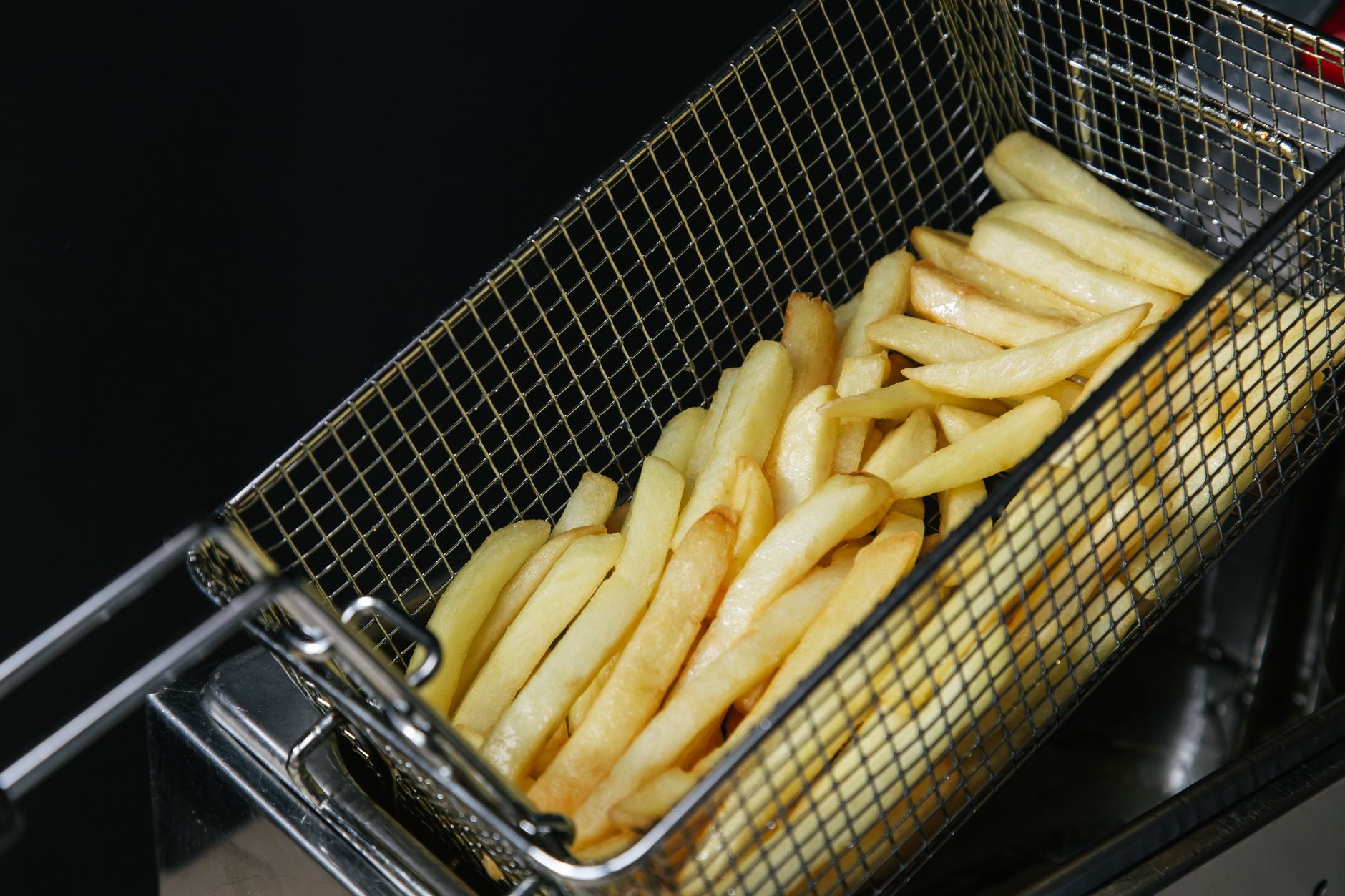 Fresh fries drain in a metal basket above hot oil in a commercial kitchen.