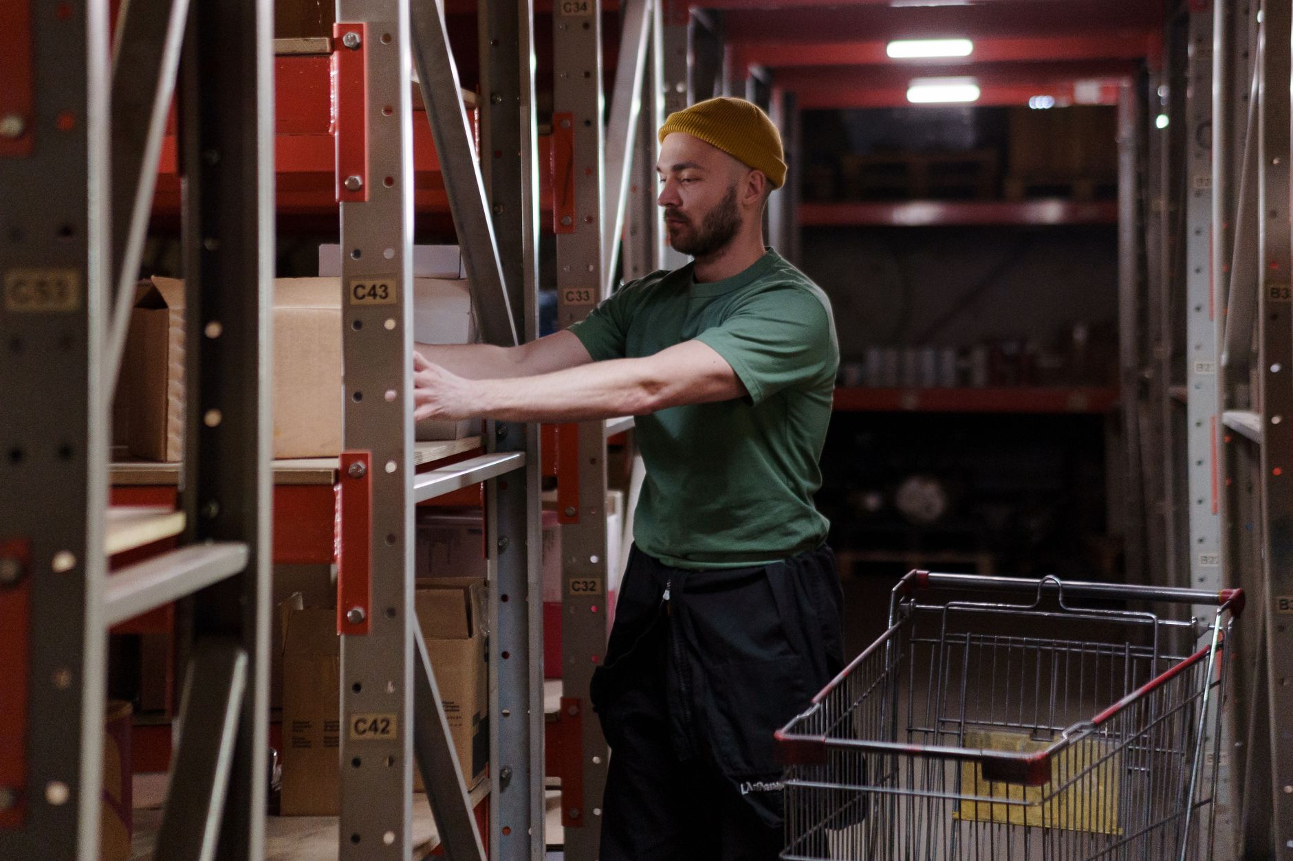 A grocery distribution worker carries a case through a warehouse aisle lined with stocked shelving.