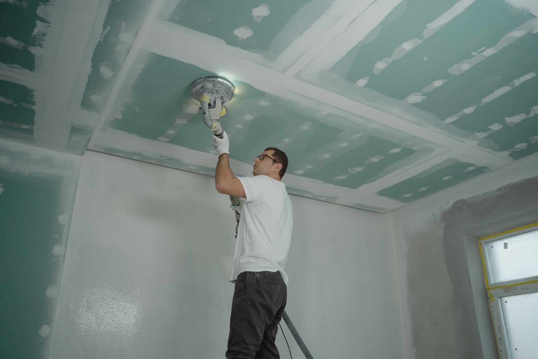 A drywall finisher sands an overhead surface inside a large commercial construction project.