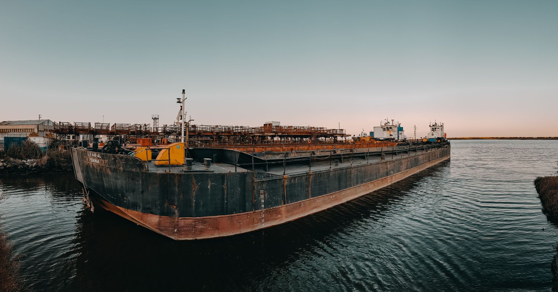 A large steel barge hull sits in a shipyard dry dock for surface preparation.