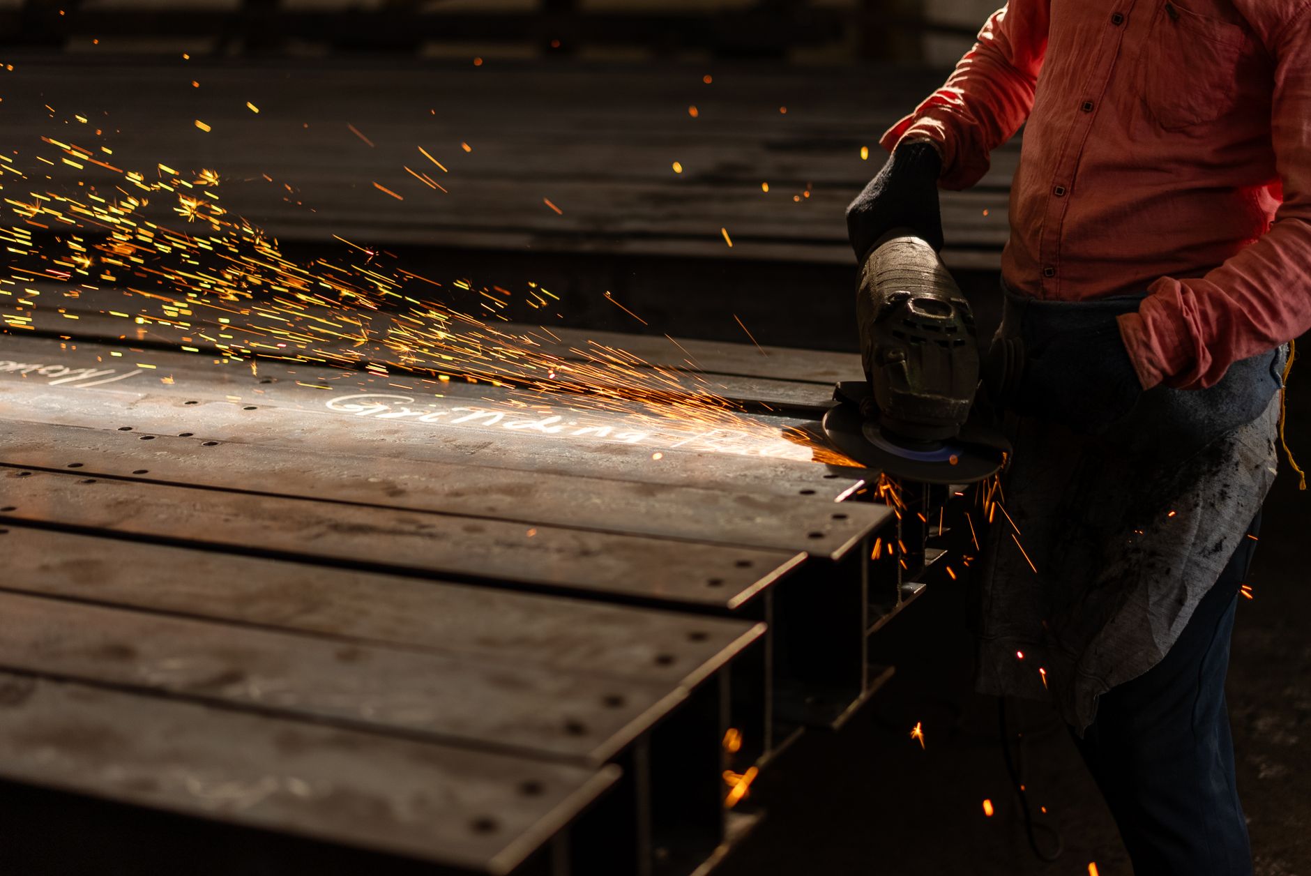 A metalworker sends sparks across a fabrication shop while grinding the edge of a steel part.