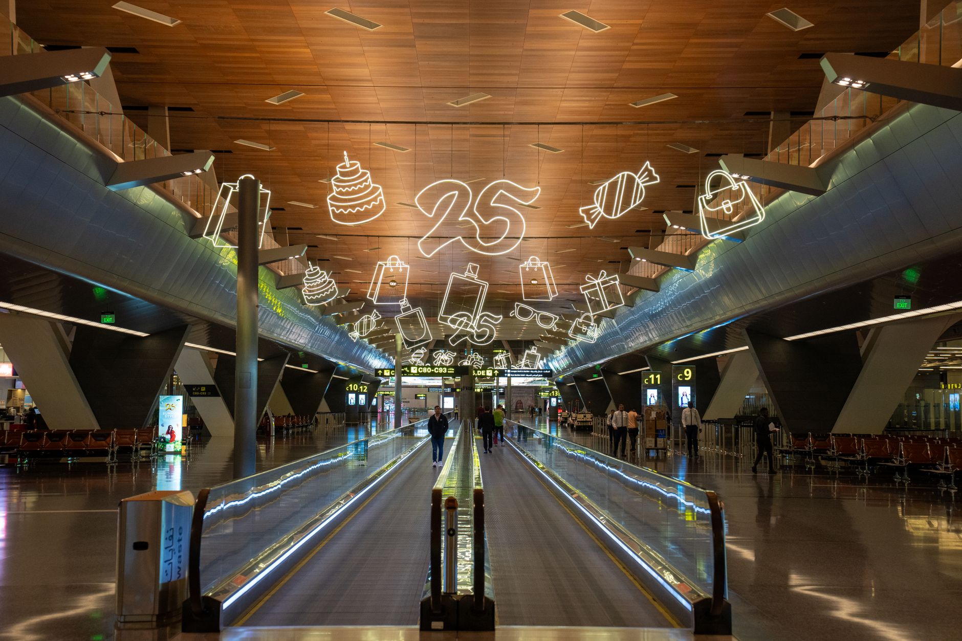 Travelers walk past brightly lit storefronts in a busy airport concourse.