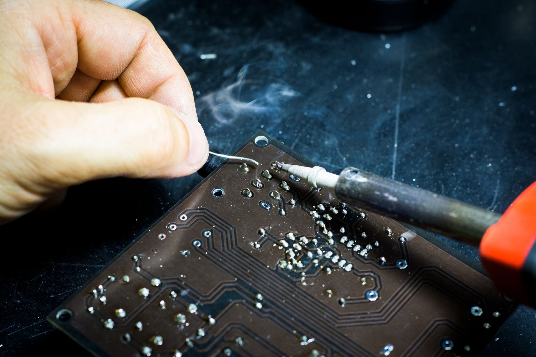 Workers assemble small electronic components at benches in a compact manufacturing facility.