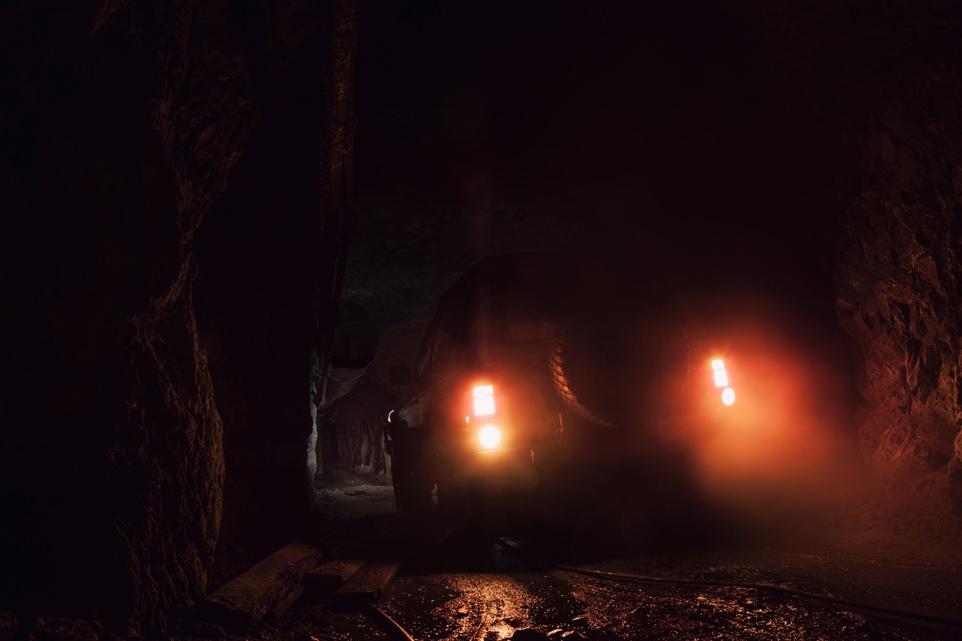 A dim underground mine tunnel with rock walls and overhead lighting, the kind of drift where loaders tram ore to passes.