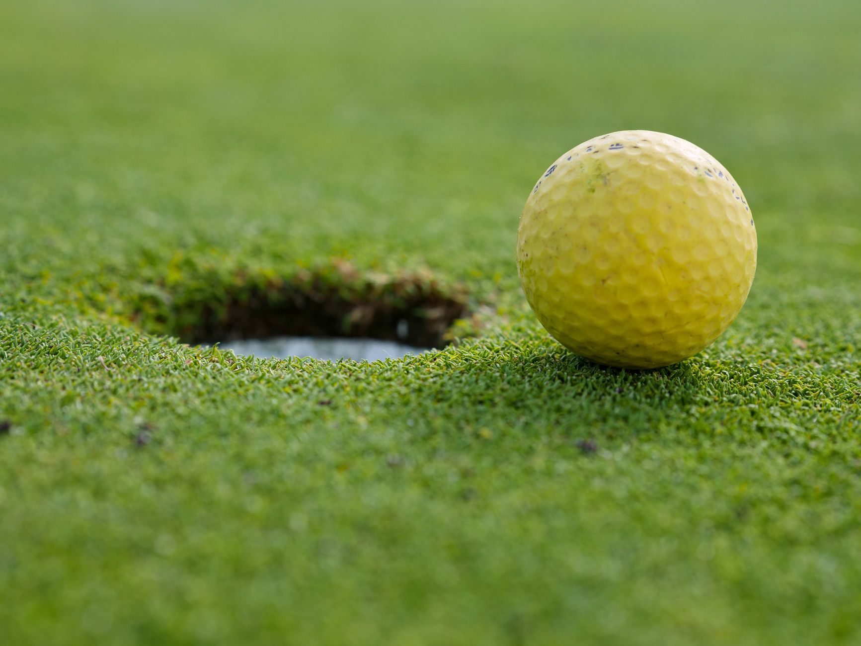 A yellow golf ball close to the hole on a grassy golf course