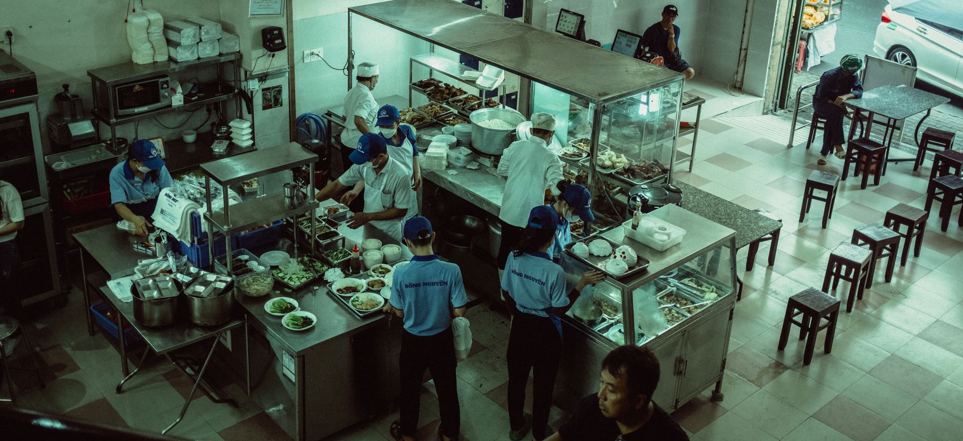 Kitchen staff in a commercial kitchen carrying stacked trays and containers past stainless steel counters.