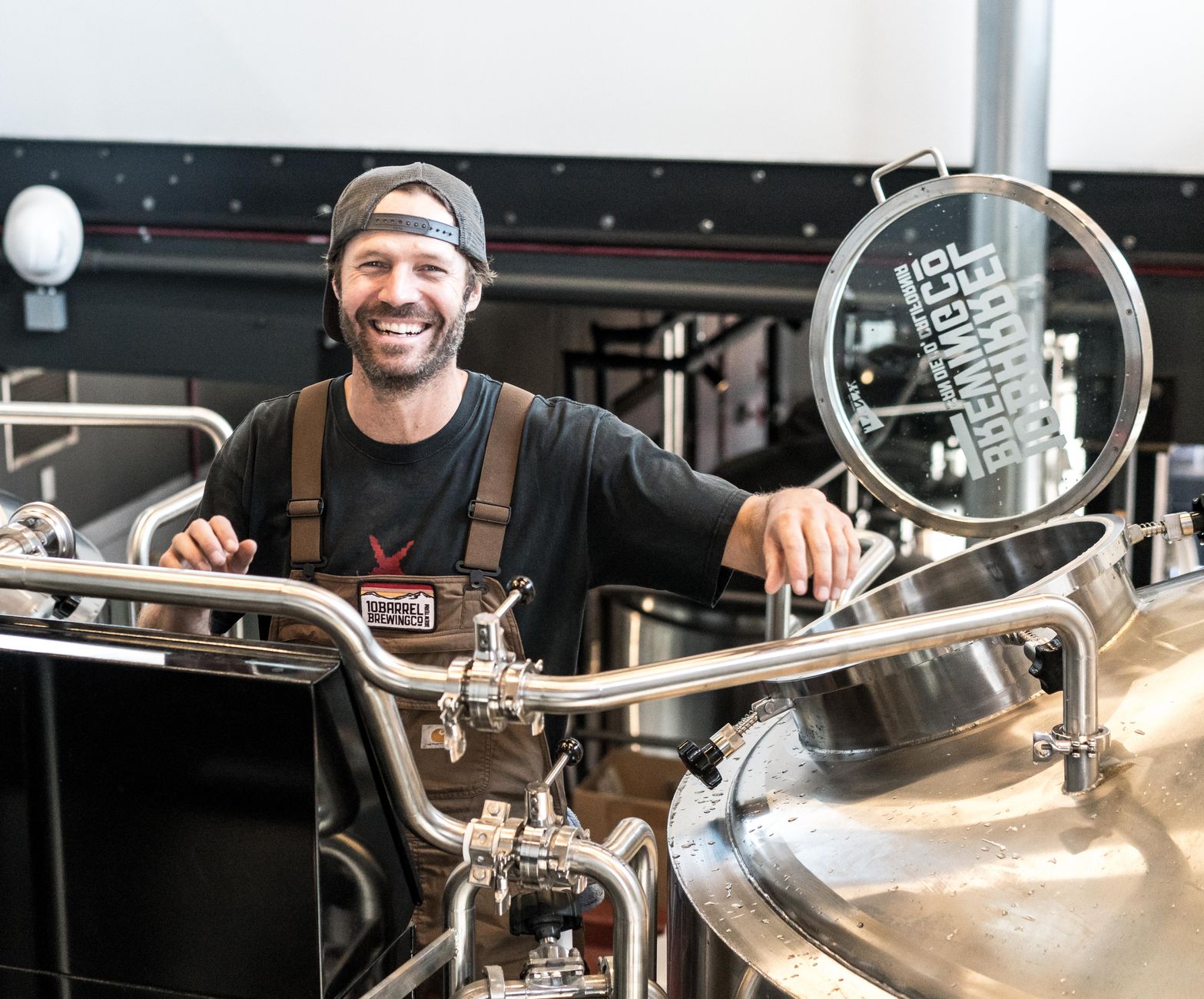 Two brewery staff members in work clothes talk beside brewing equipment.
