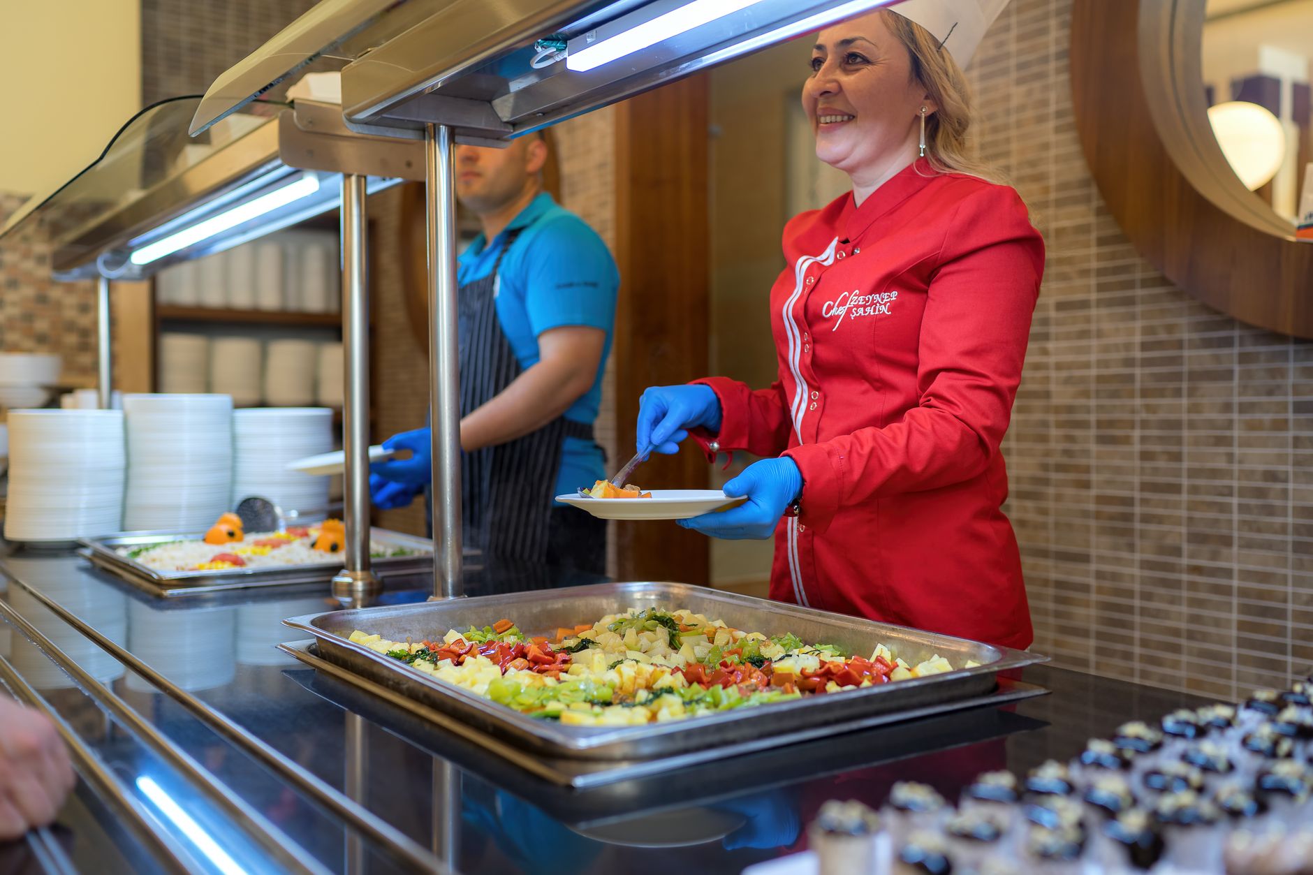 Hotel kitchen staff preparing plated food at a busy prep counter, showing the food and beverage work that staff keep doing while deliveries are handled separately.